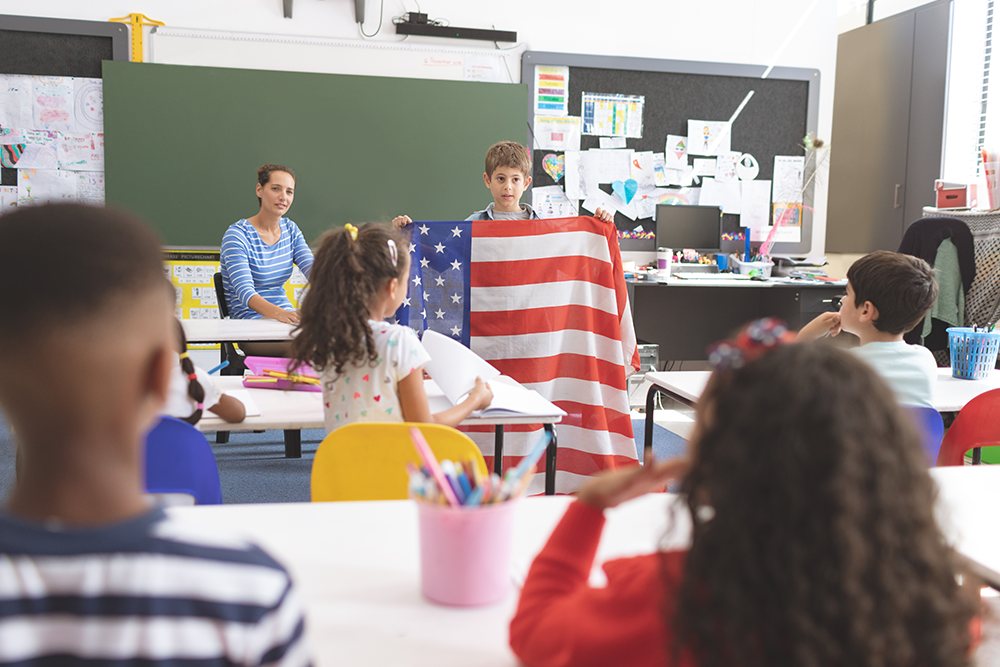 Professora explicando em sala de aula sobre a independência dos Estados Unidos enquanto aluno segura a bandeira do país