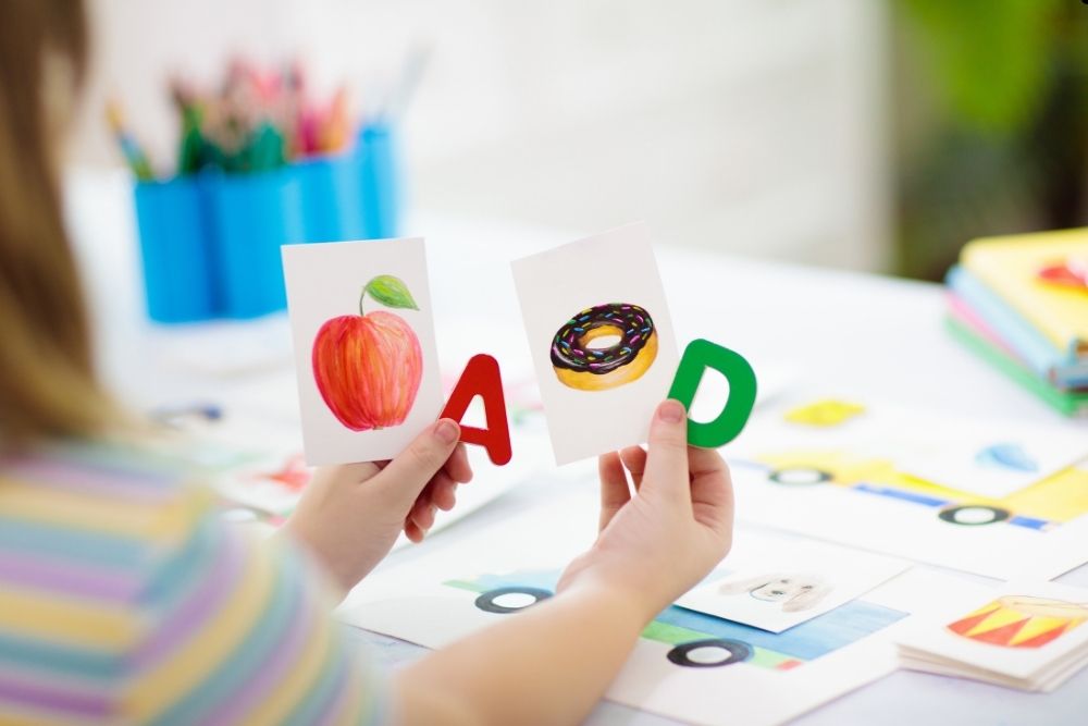 Uma pessoa segura cartas educativas com as letras “A” e “D”, cada uma acompanhada por ilustrações de um objeto. A letra “A” tem a imagem de uma maçã, enquanto a letra “D” tem a imagem de um donut.