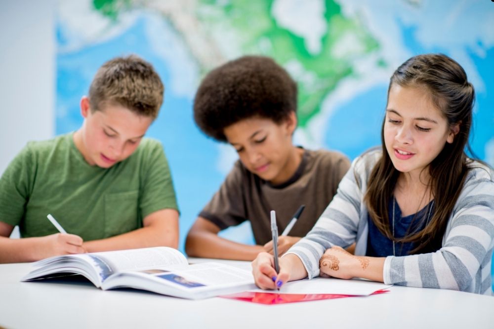 Três crianças em um ambiente escolar, praticando a escrita com um livro sobre a mesa. Ao fundo, um mapa mundi como papel de parede.