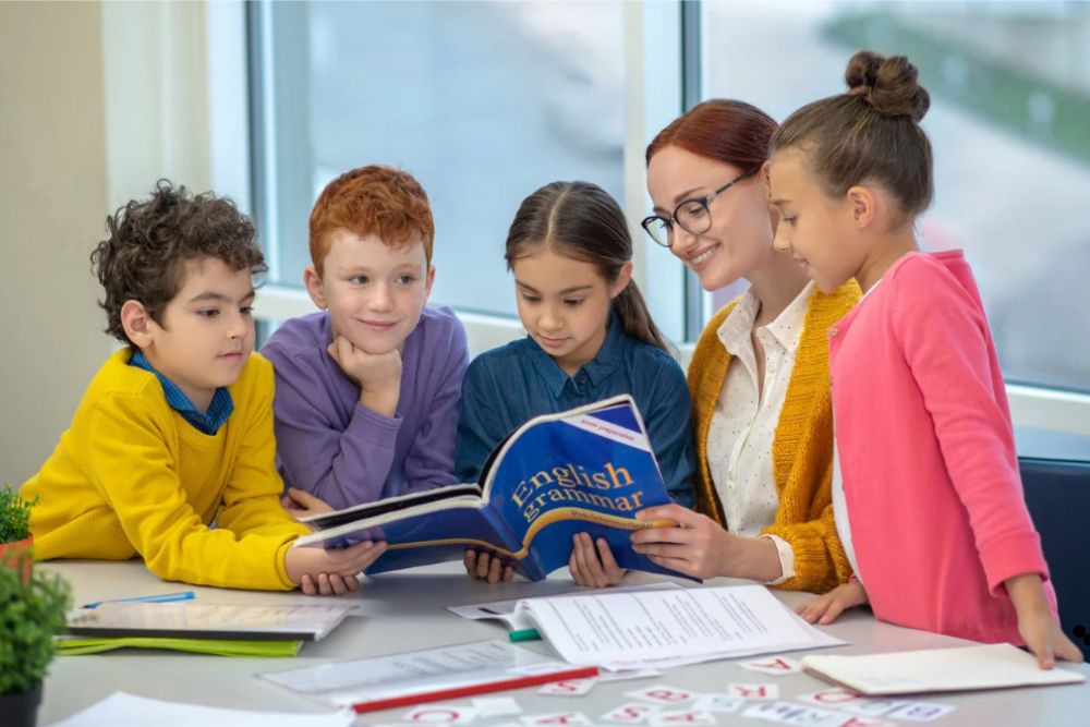 Uma professora sorridente vestindo um suéter amarelo e um grupo de quatro alunos realizando uma leitura em conjunto.